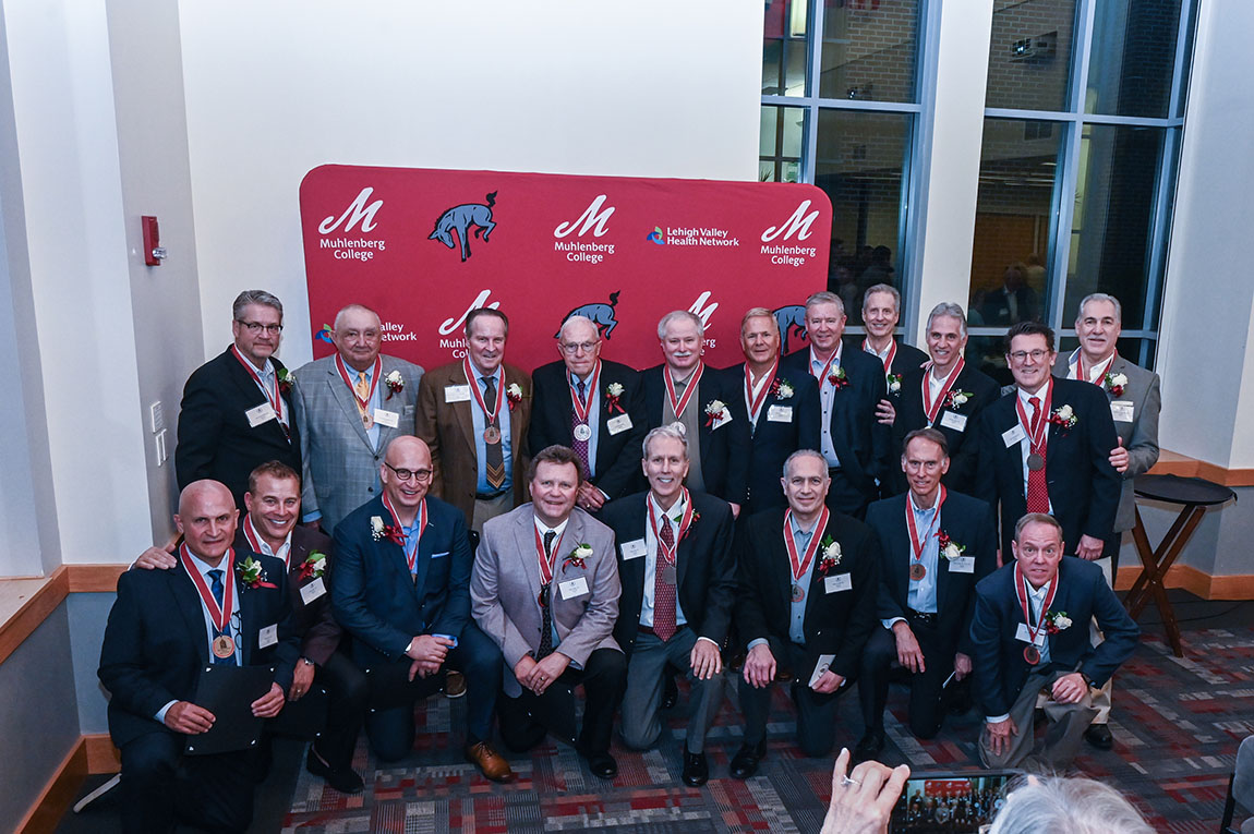 A large group of older men with medals on smile in front of a red backdrop